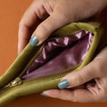 Load image into Gallery viewer, Close-up of hands opening a green zippered pouch with a purple interior on an orange background
