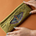 Cargar la imagen en la vista de la galería, Green clutch with embroidered moth design held by hands on an orange background
